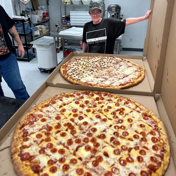 A smiling person stands behind two large pizzas, one pepperoni and one sausage, in a commercial kitchen.