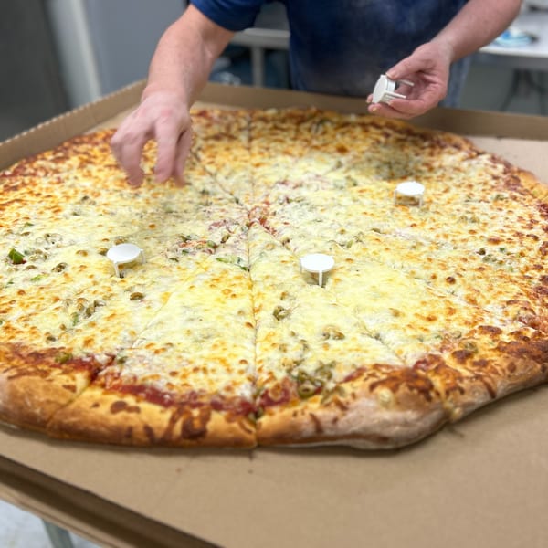 Chef's hands placing small white pizza tables on a giant, cheesy pizza in a cardboard box.